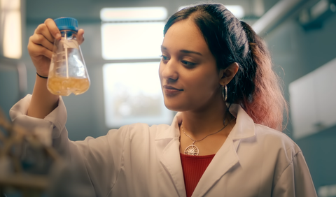 Eine junge Frau im Laborkittel betrachtet einen Glasbehälter mit einer Flüssigkeit.