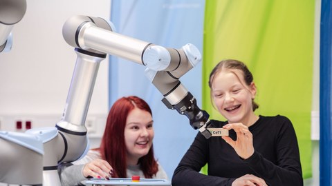 Ein Mädchen gibt einem Roboterarm einen Bauklotz. Daneben sitztz ein anderes Mädchen. Beide lachen.