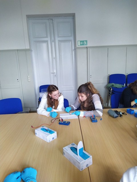 Zwei Mädchen mit Laborhandschuhen bauen etwas an einem Tisch. Auf dem Tisch liegt ein Spannungsmessgerät.