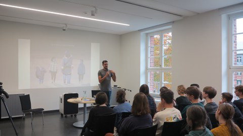 Ein Mann spricht stehend in ein Mikrofon. Vor ihm sitzen in Reihenbestuhlung ein Publikum aus jungen Erwachsenen. Im Hintergrund ist ein Bild an die Wand projiziert mit der Aufschrift: "Join us for a Lecture and Reading".