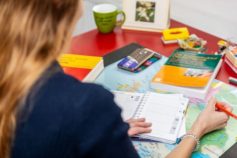 A female person is sitting at a desk. She holds a pen in her right hand. Her left hand is on a calendar in the shape of a ring binder. On the table are other objects, such as a mug, a cell phone and two books.