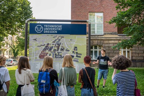 Foto. Im Vordergrund sind 6 Personen zu sehen. Sie stehen mit dem Rücken zur Kamera. Im Hintergrund ist eine große Tafel mit einem Lageplan zu sehen. Darüber steht "Technische Universität Dresden". Rechts neben der Tafel steht ein junger Mann. Er blickt in Richtung der anderen Personen.