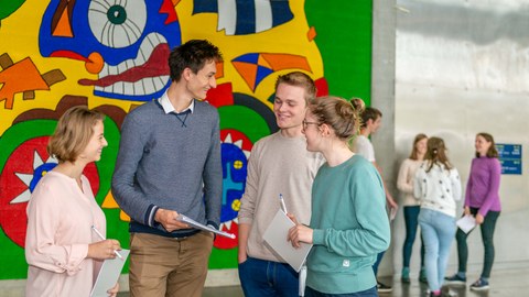 You can see a photo from the foyer of the auditorium centre. In the foreground, four young people are standing in front of a colourfully painted wall. They are talking and holding a pad and pencil in their hands. Four other people are in the background.