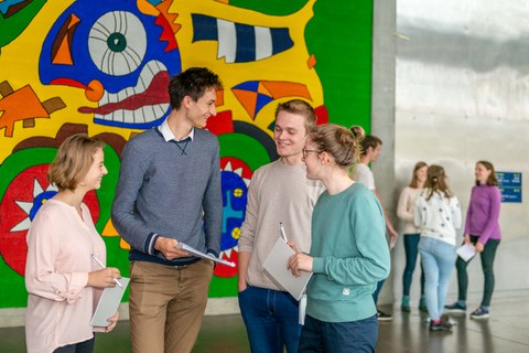 You can see a photo from the foyer of the auditorium centre. In the foreground, four young people are standing in front of a colourfully painted wall. They are talking and holding a pad and pencil in their hands. Four other people are in the background.