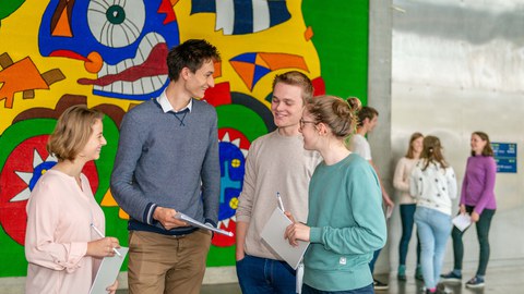 Zu sehen ist ein Foto aus dem Foyer des Hörsaalzentrums. Im Vordergrund stehen vier junge Personen vor einer bunt bemalten Wand. Sie unterhalten sich und halten Block und Stift in der Hand. Im Hintergrund sind vier weitere Personen zu erkennen.