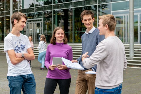 The photo shows four young people in front of the TU Dresden lecture hall centre. Three of them are each holding a pad of paper. They are talking to each other. 