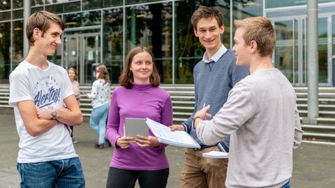 The photo shows four young people in front of the TU Dresden lecture hall centre. Three of them are each holding a pad of paper. They are talking to each other. 
