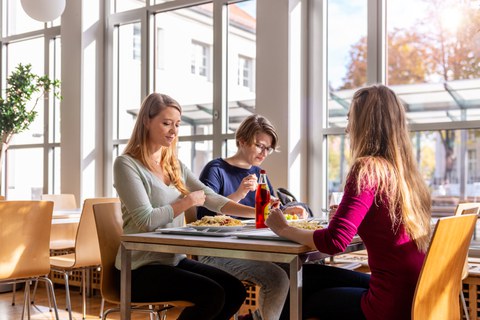 Foto von drei weiblichen Personen, die an einem Tisch sitzen. Vor ihnen stehen Tabletts mit Essen und eine Glasflasche mit einer roten Flüssigkeit. 