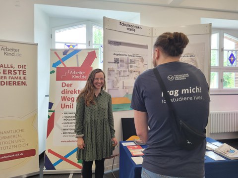 Im Vordergrund ist ein Mann von hinten zu sehen mit einem blauen T-Shirt mit der Aufschrift "Frag mich ich studiere hier". Er ist im Gespräch mit einer jungen Frau, die vor Roll-ups der Intiative ArbeiterKind.de steht. An der Seite ist der Stand der Schulkontaktstelle der TU Dresden zu sehen.