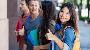 Vier junge, fröhliche Menschen stehen perspektivisch in einer Reihe. Die Studentin an erster Stelle lächelt in die Kamera und hält den gestreckten Daumen hoch. 