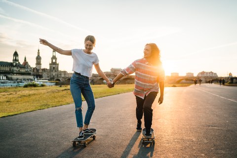 Eine junge Frau und ein junger Mann fahren mit dem Skateboard am Elbufer und halten sich an den Händen. Im Hintergrund befindet sich die Skyline von Dresden.