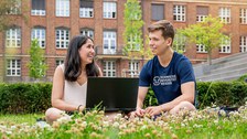 Bild von zwei Studenten, die lachend auf der Wiese zusammen am Laptop arbeiten.