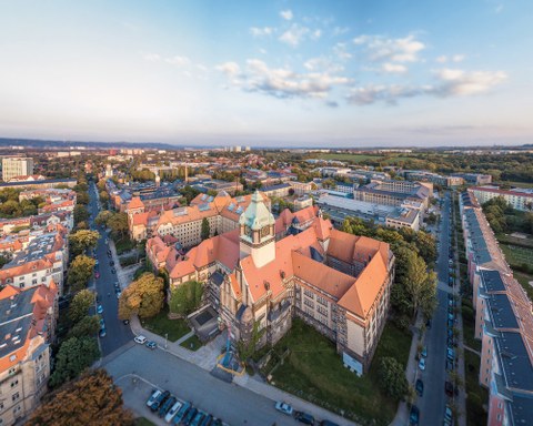 Das Foto zeigt eine Luftaufnahme des Georg-Schumann-Baus der TU Dresden. Das Gebäude wirkt wie eine Art Burg. Besonders auffällig sind der große Wehrturm und die spitzen Dächer. 