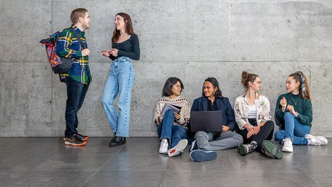 Eine Gruppe Studierender, teilweise stehend, teilweise sitzend, vor einer Betonwand