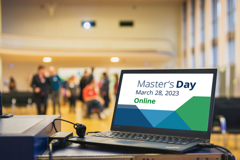 The photo shows an event hall. In the foreground you can see a laptop on the speaker's desk. In the laptop is a picture with colored tiles and the lettering MasterTag March 28, 2023 Online.