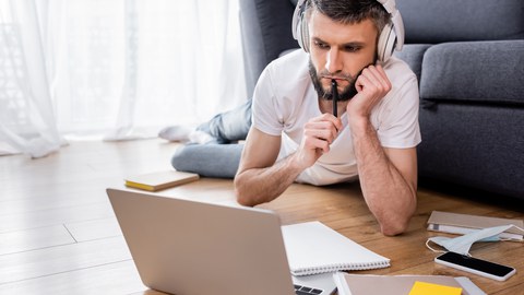 junger Mann mit Kopfhörern liegt auf dem Boden und schaut sehr angestrengt auf seinen Laptop