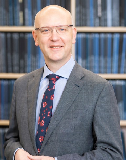 Auf dem Hochkant-Foto ist ein lächelner Mann im grauen Anzug mit Glatze und Brille vor einer blaufarbigen Bücherwand zu sehen. Er trägt eine dunkelblaue Krawatte mit roten Blüten.
