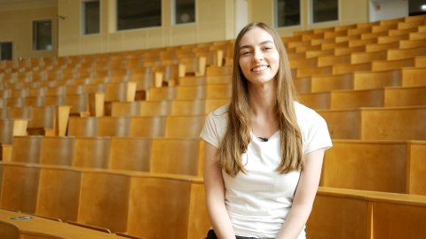 Studentin Rebecca sitzt im leeren Hörsaal und schaut in die Kamera.