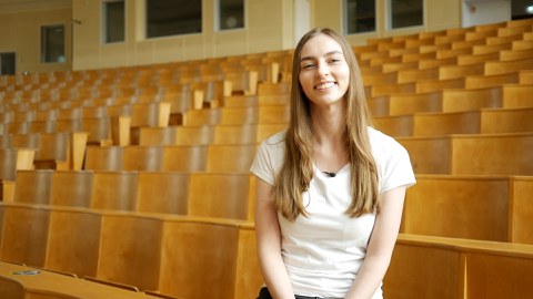 Studentin Rebecca sitzt im leeren Hörsaal und schaut in die Kamera.