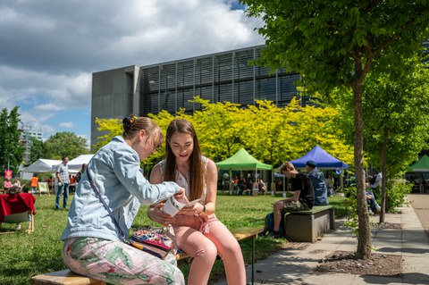 Zwei junge Frauen schauen hinter dem HSZ der TUD in einen Flyer. Es ist Sommer.