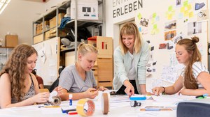 Drei junge Frauen sitzen am Tisch und eine Frau steht am Tisch. Auf dem Tisch liegen viel Papier, Schere, Leim und Stifte. Die Frauen basteln aus dem Papier Modelle.