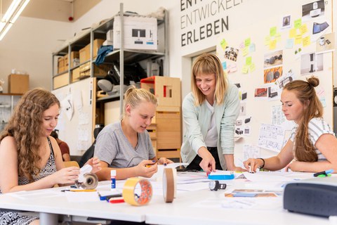 Drei junge Frauen sitzen am Tisch und eine Frau steht am Tisch. Auf dem Tisch liegen viel Papier, Schere, Leim und Stifte. Die Frauen basteln aus dem Papier Modelle.