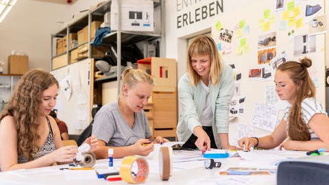 Drei junge Frauen sitzen am Tisch und eine Frau steht am Tisch. Auf dem Tisch liegen viel Papier, Schere, Leim und Stifte. Die Frauen basteln aus dem Papier Modelle.