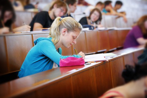 Das Foto zeigt einen Hörsaal voller Studierender. Der Fokus liegt auf einer Studentin. Sie notiert mit einem Bleistift gerade etwas in ihren Unterlagen.