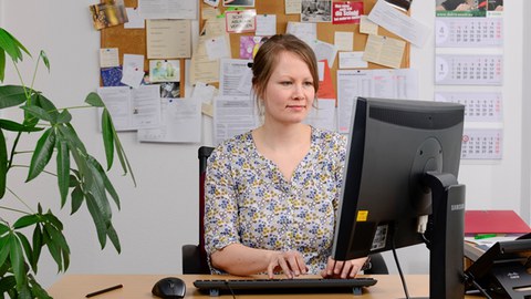 Das Bild zeigt ein Foto von einer Studienberaterin. Sie sitzt an ihrem Arbeitsplatz vor ihrem PC und tippt auf der Tastatur. 