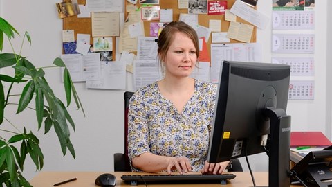 The image shows a photo of a student counsellor. She is sitting at her workplace in front of her PC and typing on the keyboard. 