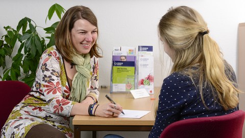 Es ist ein Foto zu sehen, auf dem zwei Personen abgebildet sind. Sie sitzen sich gegenüber an einem Tisch. Die Person links im Bild ist eine Studienberaterin, welche sich Notizen macht. Die zweite Person ist nur von hinten zu erkennen.