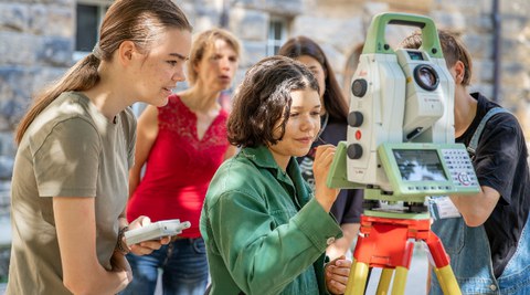 There is a photo outside an older building. Five people are inside. Two students are operating a surveying device. The other people are watching them from the background.