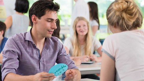 Es ist eine Momentaufnahme aus einem Workshop in einem hellen Seminarraum zu sehen. Einige Studierende sitzen in Zweiergruppen an jeweils einem Tisch. Andere stehen im Hintergrund mit einer Studienberaterin vor einem Flipchart.