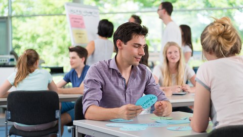 Es ist eine Momentaufnahme aus einem Workshop in einem hellen Seminarraum zu sehen. Einige Studierende sitzen in Zweiergruppen an jeweils einem Tisch. Andere stehen im Hintergrund mit einer Studienberaterin vor einem Flipchart.