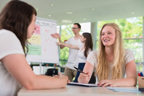 Es ist eine Momentaufnahme aus einem Workshop in einem hellen Seminarraum zu sehen. Zwei junge Frauen sitzen im Vordergrund an einem Tisch und unterhalten sich. Zwei andere junge Menschen stehen im Hintergrund vor einem Flipchart.