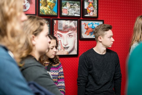 You can see a photo of some young people in front of a red wall. They are all looking in one direction. On the wall hang colorful photos with different motifs.