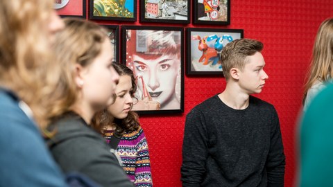 You can see a photo of some young people in front of a red wall. They are all looking in one direction. On the wall hang colorful photos with different motifs.