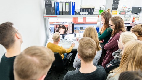 Auf dem Foto ist eine Gruppe Jugendlicher in einem Büro zu sehen. Sie stehen in einem Halbkreis um einen Arbeitsplatz mit Monitor und Tastatur herum. An diesem Platz sitzt ein Mann. Ihre Blicke richten sich auf den Monitor.
