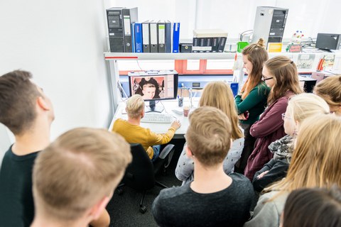 Auf dem Foto ist eine Gruppe Jugendlicher in einem Büro zu sehen. Sie stehen in einem Halbkreis um einen Arbeitsplatz mit Monitor und Tastatur herum. An diesem Platz sitzt ein Mann. Ihre Blicke richten sich auf den Monitor.