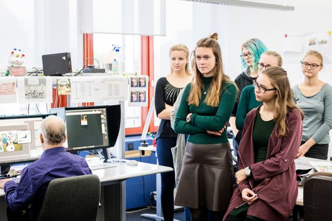 Auf diesem Foto sind einige Teilnehmerinnen der Herbstuni zu sehen. Sie stehen in einem Büro.  Links Im Bild ist ein Mann vor zwei Monitoren zu erkennen, welcher ein Bild bearbeitet.