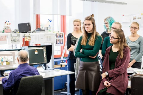 This photo shows some participants of the Autumn University. They are standing in an office.  On the left of the picture, a man can be seen in front of two monitors editing a picture.