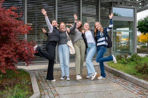 Es sind fünf Teilnehmer:innen der Herbstuniversität zu sehen, die in die Luft springen und fröhlich schauen. Sie stehen vor dem Gebäude der AOK PLUS, einem Projektpartner der Herbstuniversität.