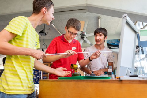 Es ist ein Foto von drei jungen Männern in einer Werkstatt zu sehen. Sie stehen neben einem Tisch und halten ein langes, dünnes Rohr in der Hand. Auf dem Tisch sind ein Schraubstock und kleine Gewichte zu erkennen.
