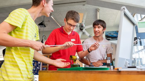 Es ist ein Foto von drei jungen Männern in einer Werkstatt zu sehen. Sie stehen neben einem Tisch und halten ein langes, dünnes Rohr in der Hand. Auf dem Tisch sind ein Schraubstock und kleine Gewichte zu erkennen.