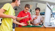 Es ist ein Foto von drei jungen Männern in einer Werkstatt zu sehen. Sie stehen neben einem Tisch und halten ein langes, dünnes Rohr in der Hand. Auf dem Tisch sind ein Schraubstock und kleine Gewichte zu erkennen.