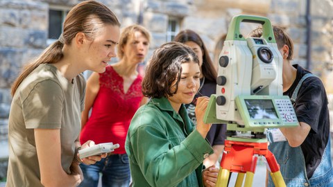 A group of female students with supervisors are standing in front of a surveying equipment outside. A female student is working on the surveying device.