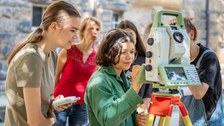 Eine Gruppe von Schülerinnen mit Betreuer:innen stehen vor einem Vermessunsgerät im Freien. Eine Schülerin arbeitet am Vermessungsgerät.