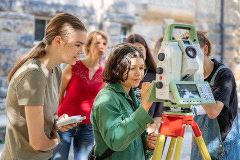 A group of female students with supervisors are standing in front of a surveying equipment outside. A female student is working on the surveying device.