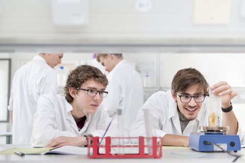 Es ist ein Foto zu sehen. Vier Personen befinden sich in einem Labor. Sie tragen weiße Kittel. Zwei männliche Studieninteressierte sitzen im Vordergrund an einem Tisch und experimentieren. Im Hintergrund stehen ebenfalls zwei Jugendliche.