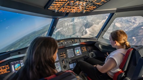 Zwei Schülerinnen sitzen im Cockpit eines Flugsimulators und steuern das Flugzeug. Das Flugzeug befindet sich in Schräglage und überfliegt Gebirge.